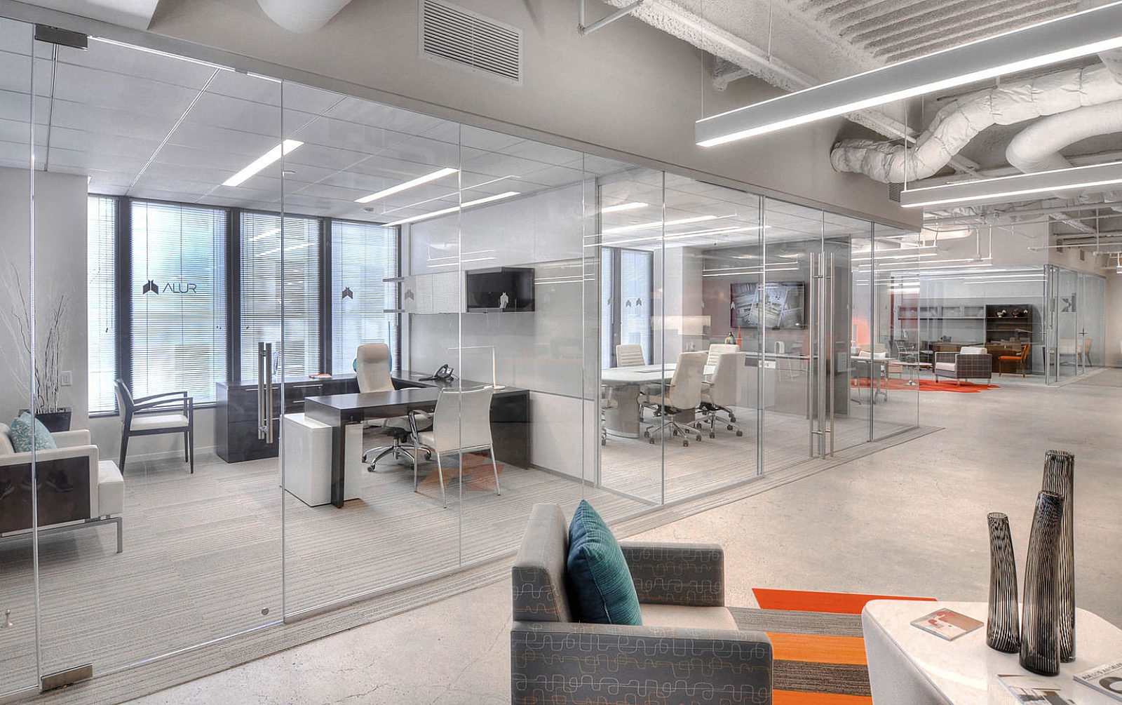 Bright space with a row of glass-front offices; clear glass with silver trim; U-shape desk in one office and conference setting in the other.