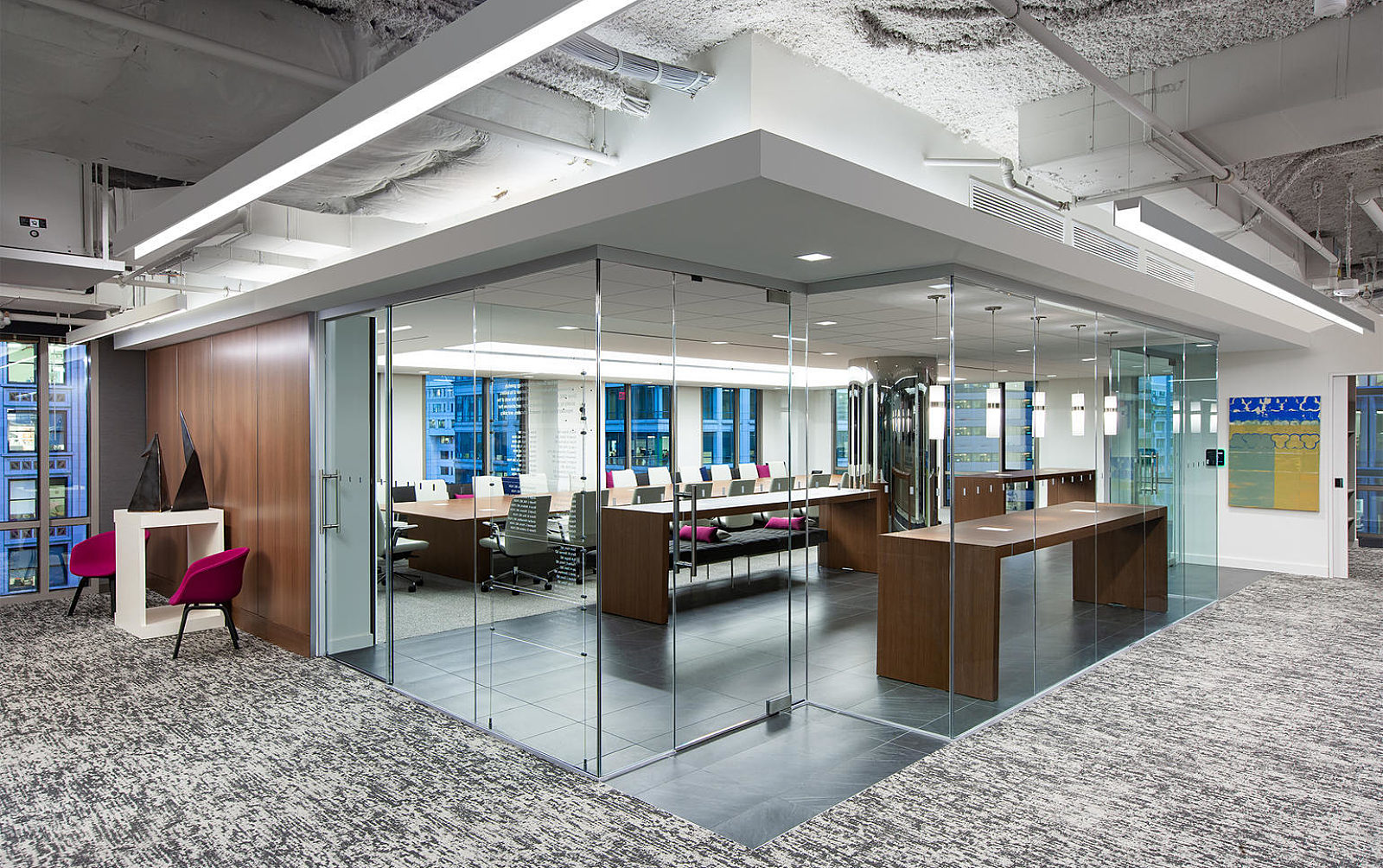 Corner perspective of a center room with full glass walls and doors; dark wood collaborative and conference tables inside room. Pops of magenta throughout