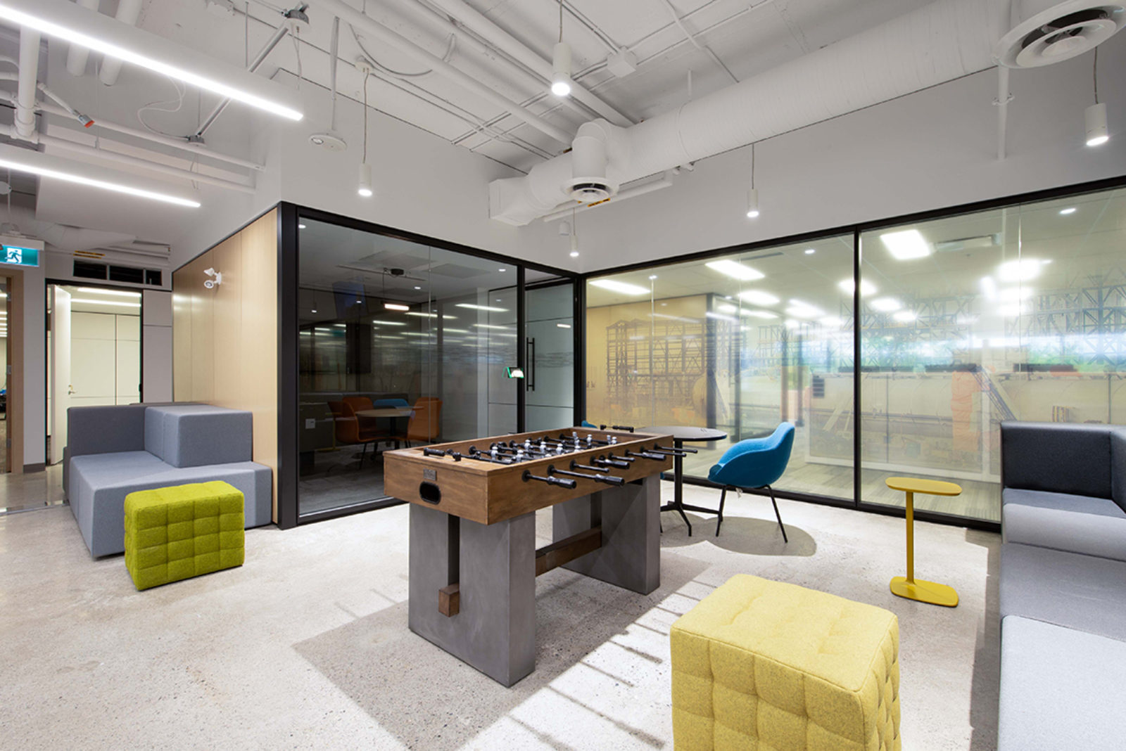 Industrial room with concrete floor, exposed ceilings painted white. Foosball table, lounge pieces and DIRTT modular room create a fun energetic space