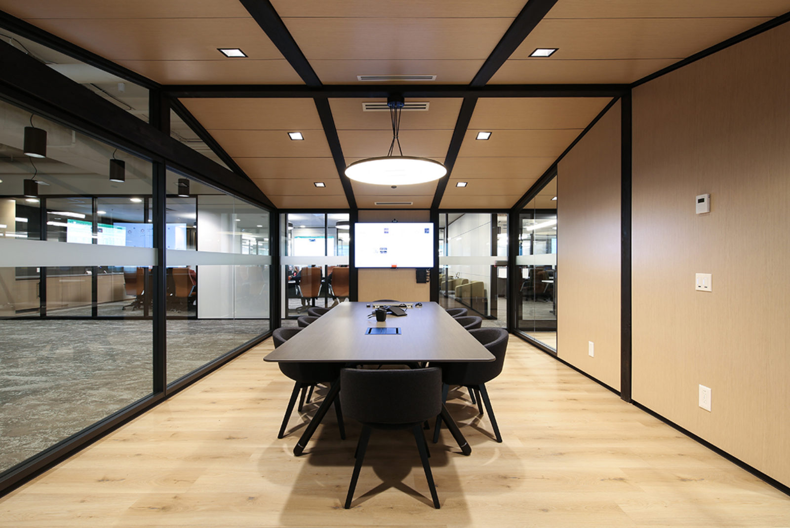 Inside of a DIRTT modular room. Conference setting with light hard floors, black beams, wooden panels and glass. Table and chairs are in the center.