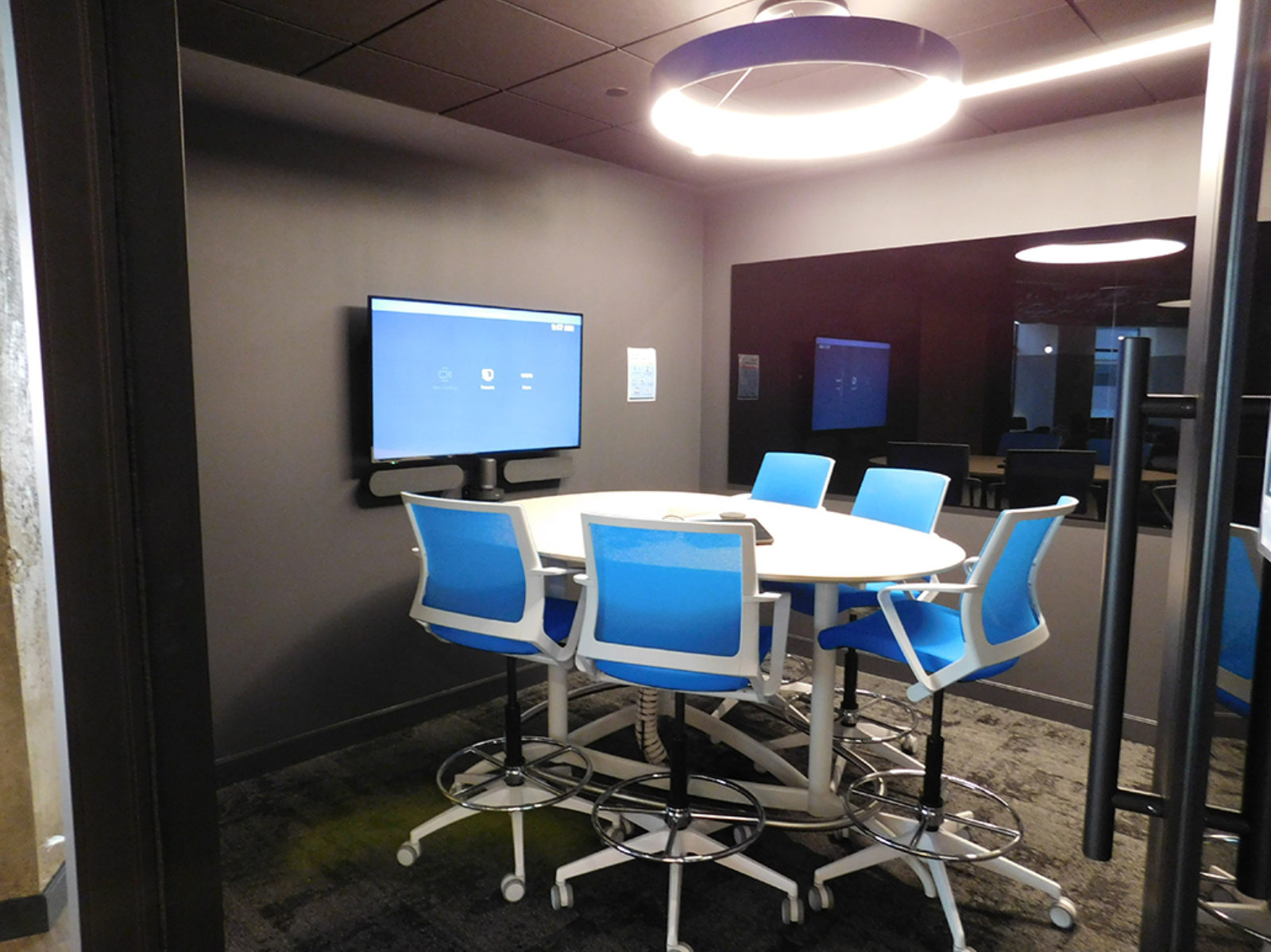 Collaborative space with dark grey walls, bright blue mesh stools, bar-height table with TV screen on wall