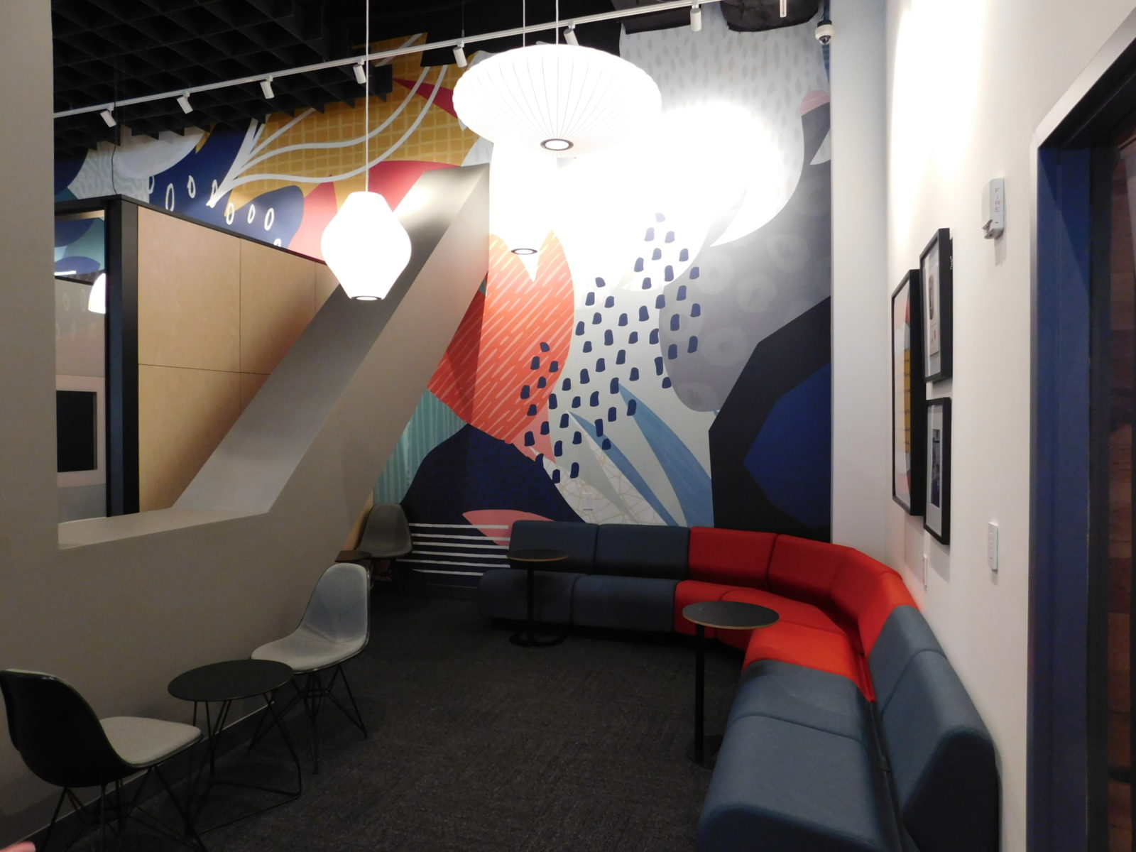 Lounge area with curved-shaped Herman Miller Chadwick sofa, dark blue and red fabric; some small laptop tables and Eames side chairs with grey fabric.