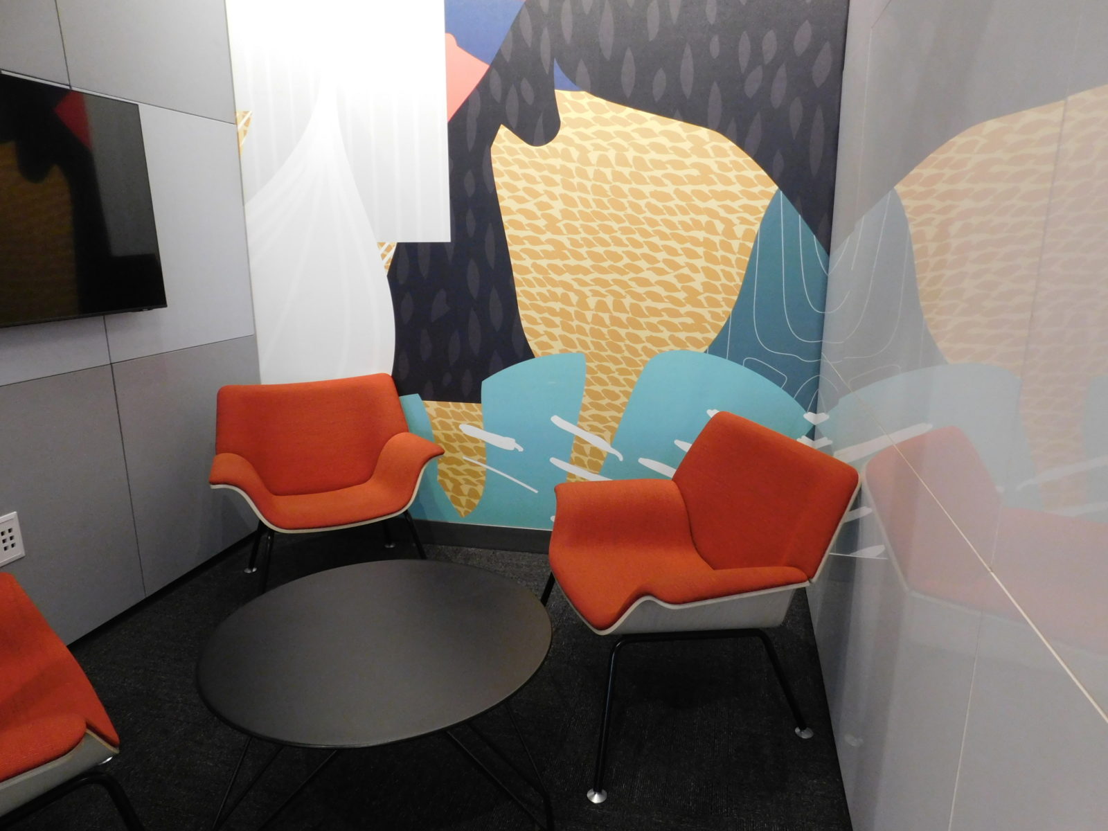 Inside of a DIRTT room with 3 red Herman Miller Swoop chairs and a black occasional table in the middle. TV on left wall, graphic wall paper on back wall.
