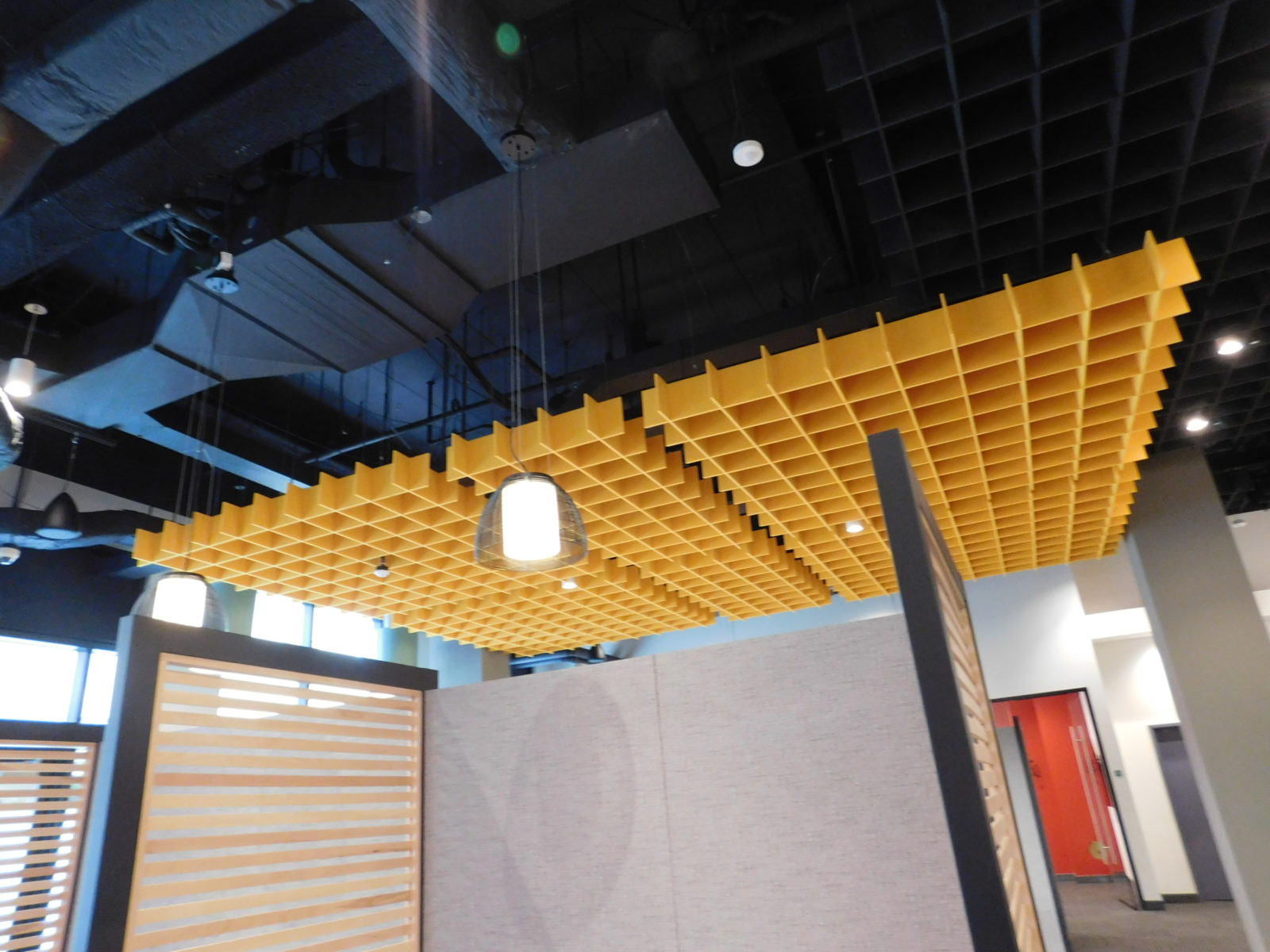 View of a black-painted ceiling with hanging yellow acoustical ceiling clouds. The acoustic panels are hanging over a Herman Miller Overlay unit.