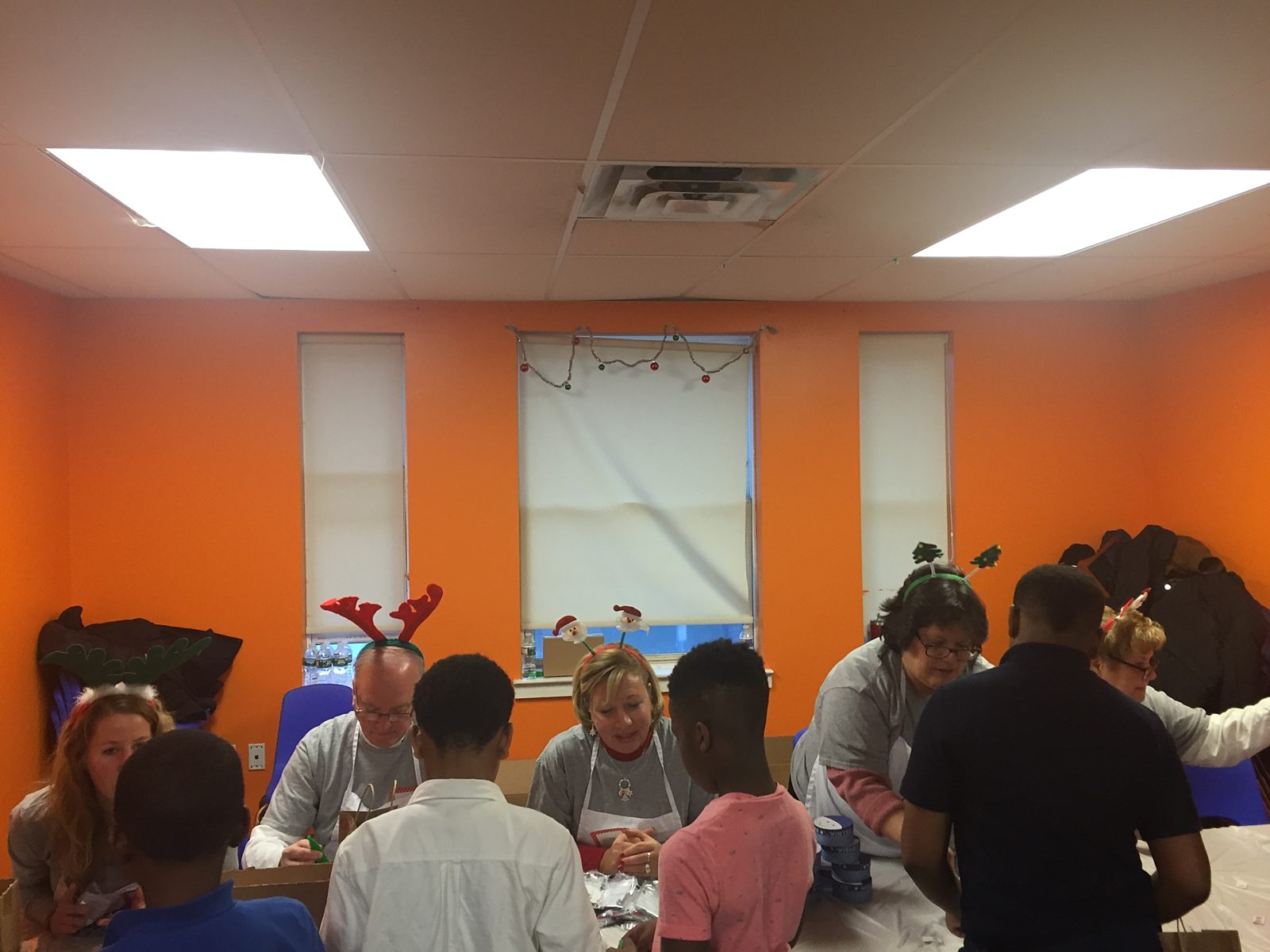 Orange wall in the background; a table of Premier employees helping children make holiday craft gifts for their family.
