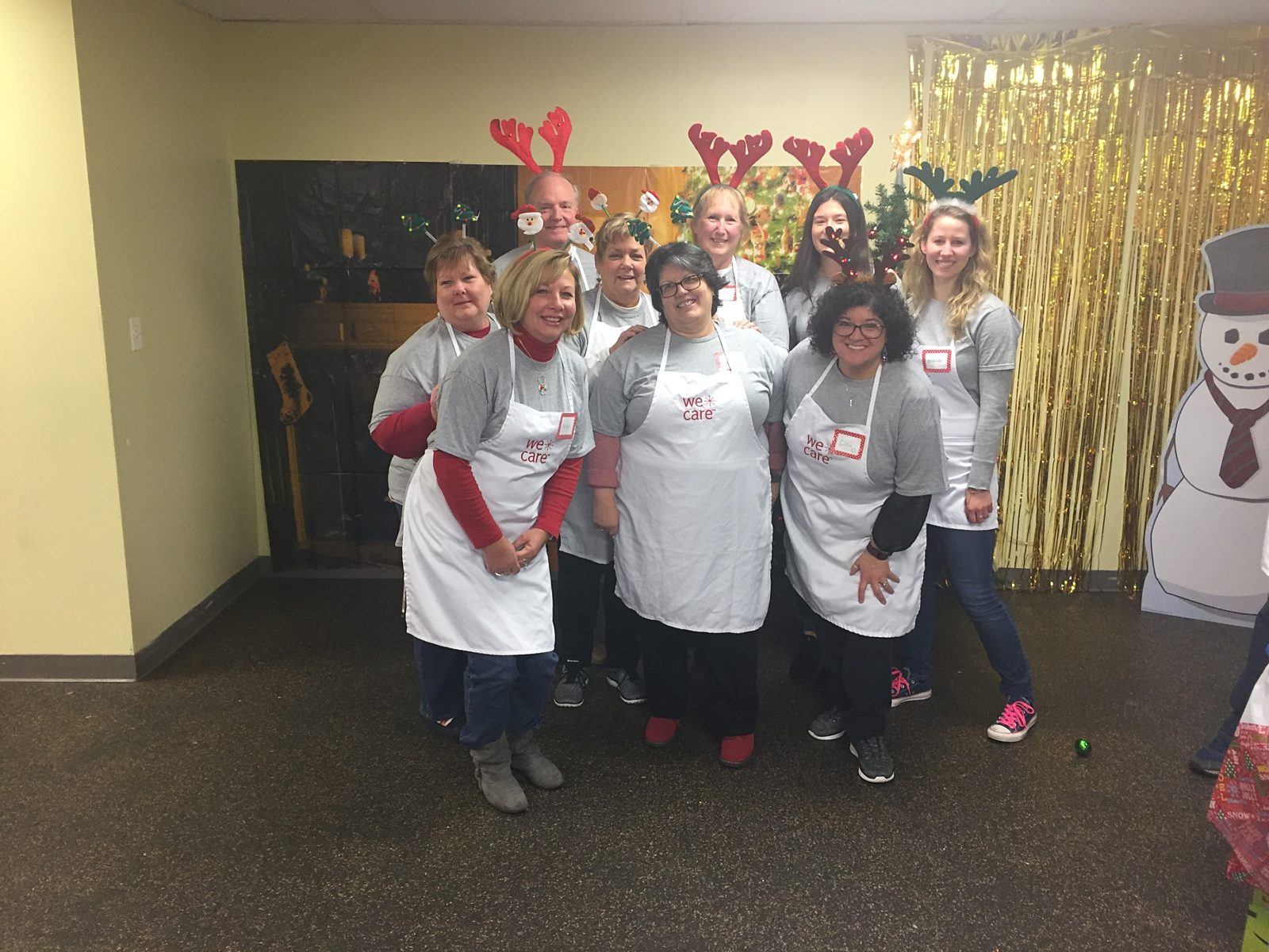 Group picture of nine Premier employees wearing "We Care aprons" and holiday reindeer headbands. There is a gold backdrop hanging behind.