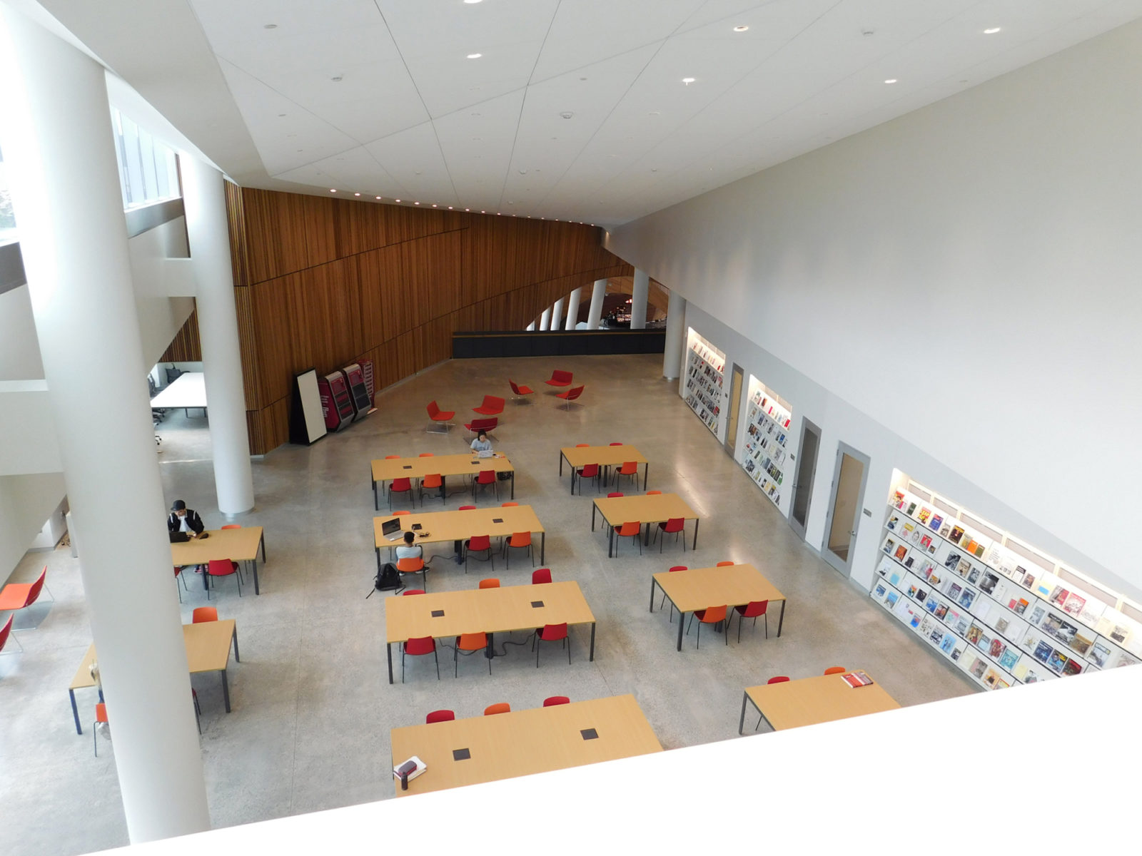 View of open area from above. There are several tables and chairs set up in the space and there is a wall of books on the right side.