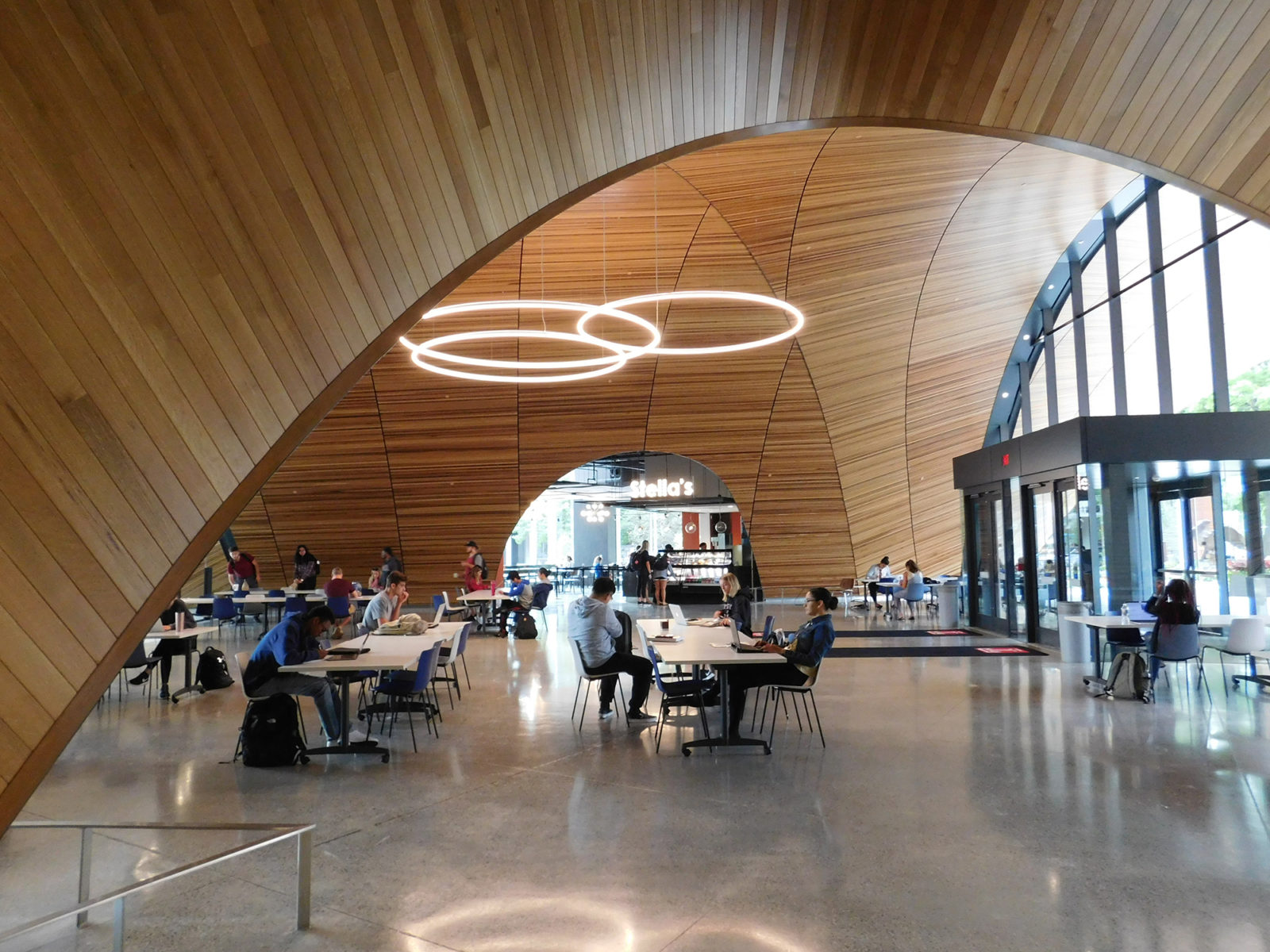View of open cafe area showing wooden architectural arches and ceilings. Featured accent lighting hangs over clusters of tables and chairs with people.