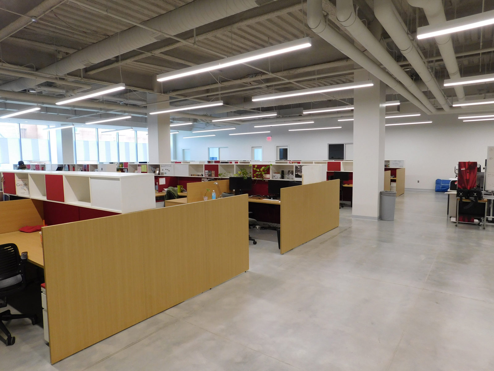 Open office space with workstations - light woodgrain surfaces and gallery panels, white and red overheads. Exposed ceilings and concrete floors.