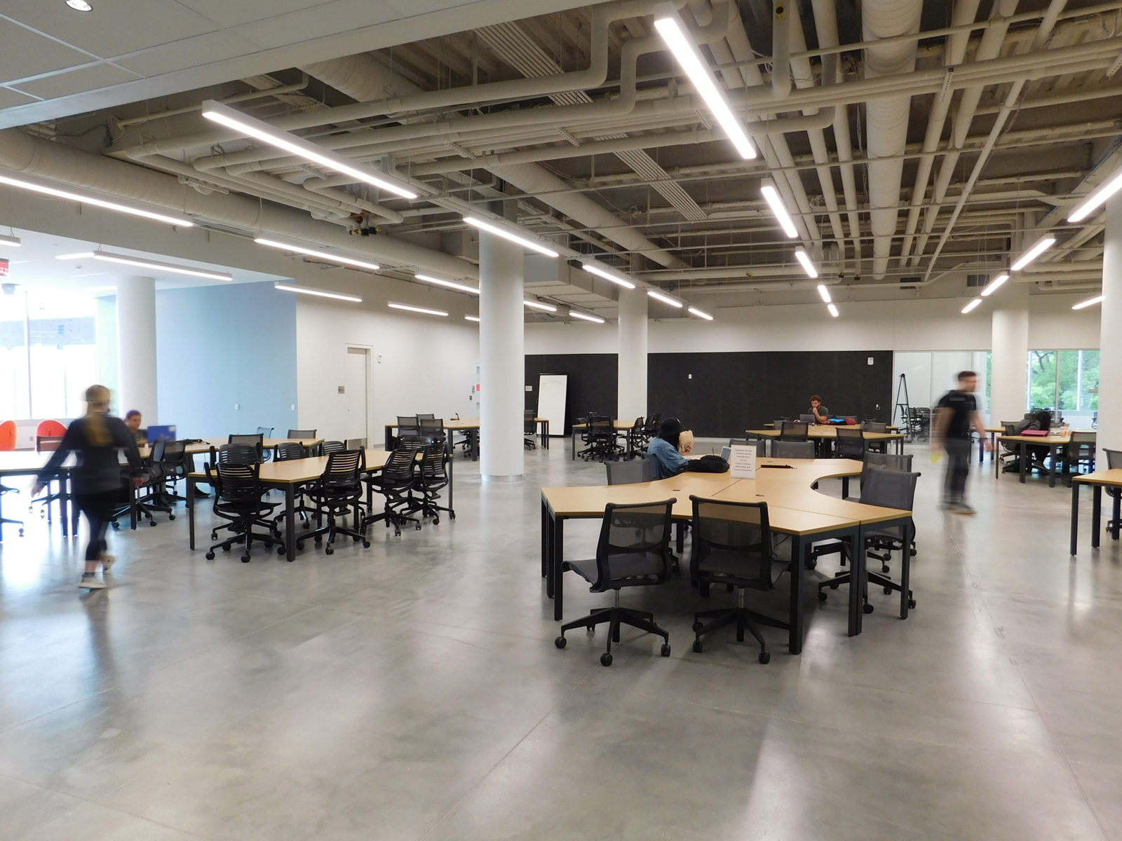 Open space with 120-degree workstations for students, including black mesh back task chairs. Exposed ceilings and concrete floors.