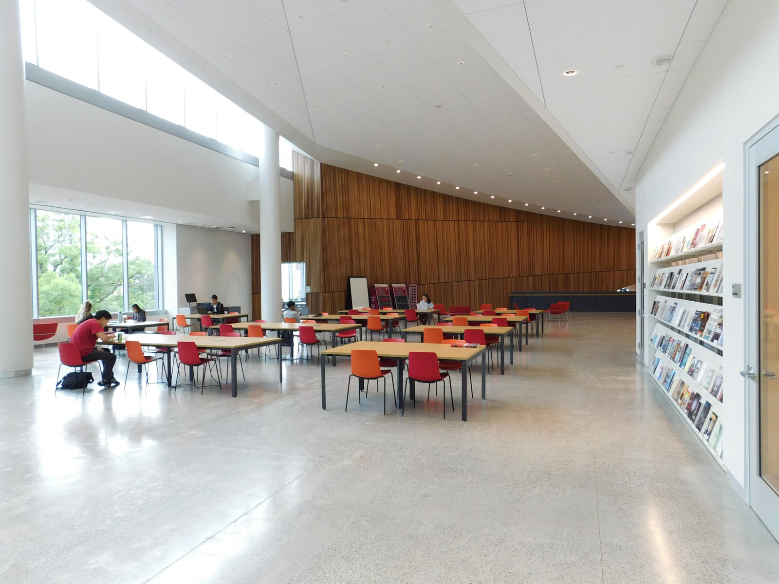 Open space with woodgrain tables and red and orange chairs. Concrete floors, wood accent wall in the background and a wall of books along the right.