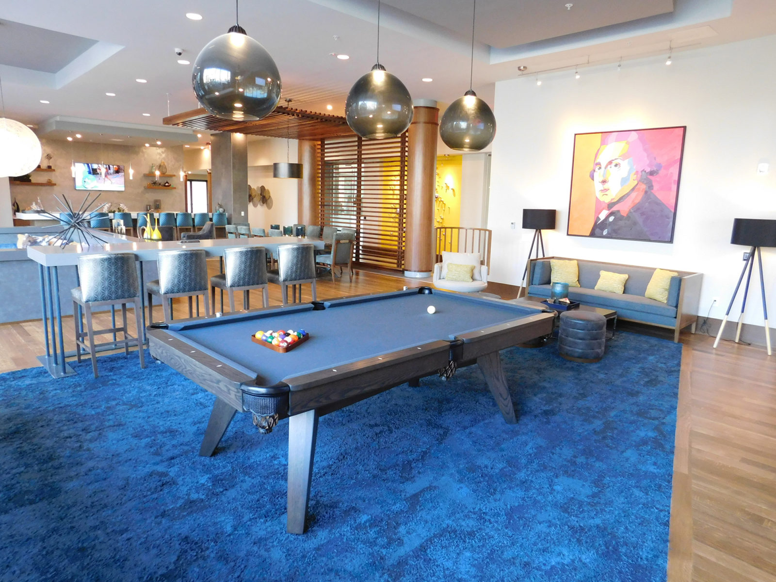 Common area with a pool table under 3 pendant lights and sitting on a blue carpet. There is a set of 4 stools behind at a seating area.