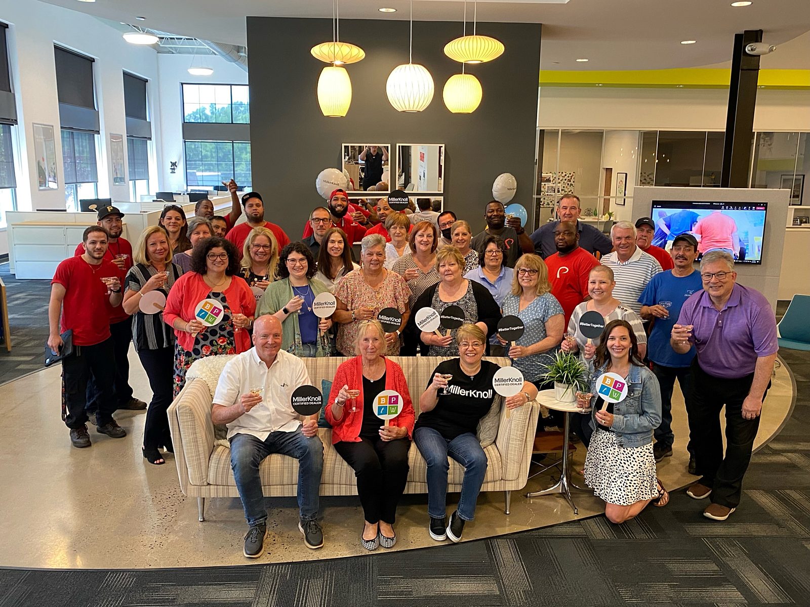Group shot of our employees holding a glass of champagne and a round paper sign (Best Places to Work, MillerKnoll Certified Dealer, #BetterToghether)