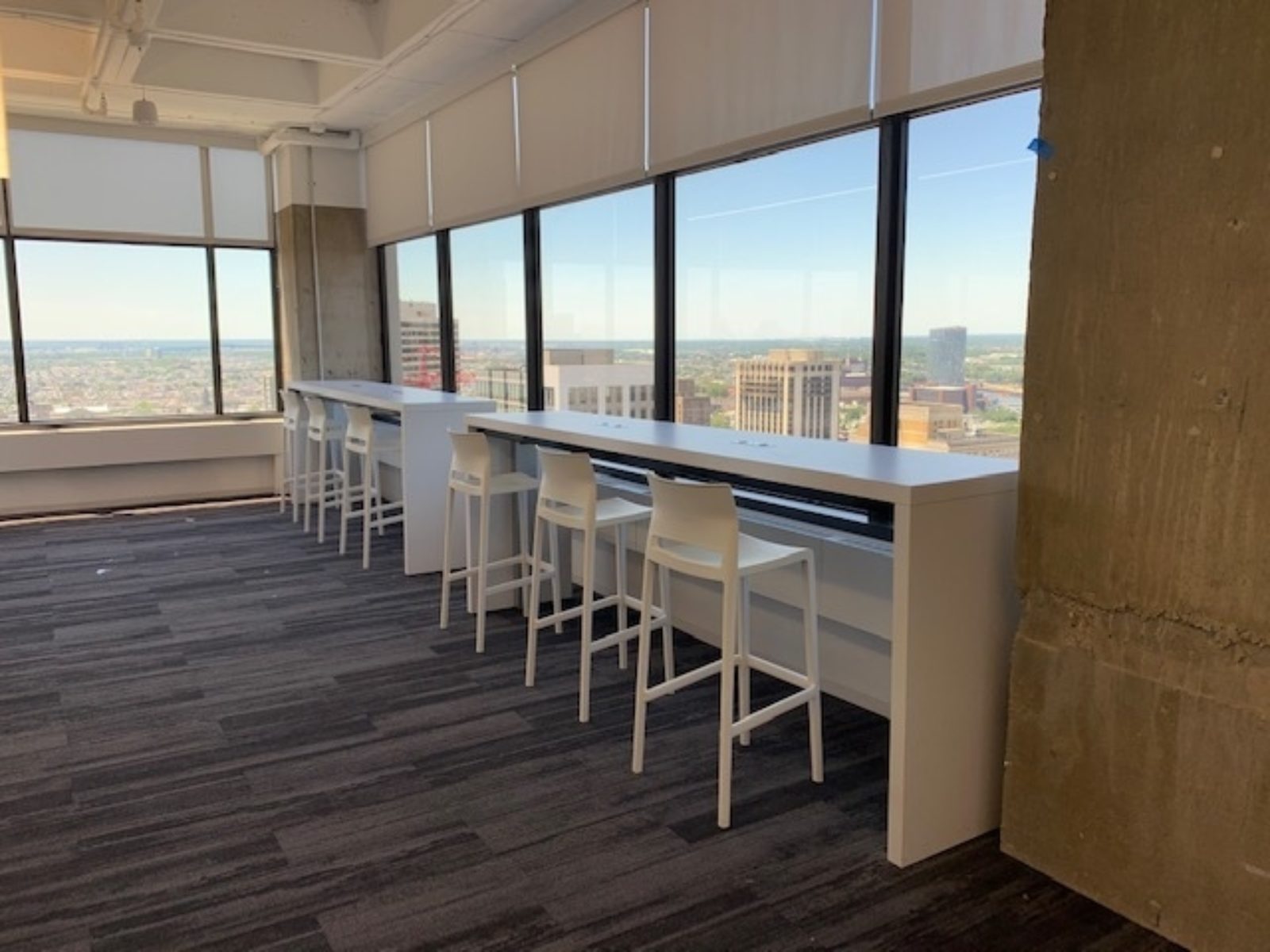 Bar-height white tables with white barstools; tables are along a wall of windows so the chairs are facing out to the view of the city.