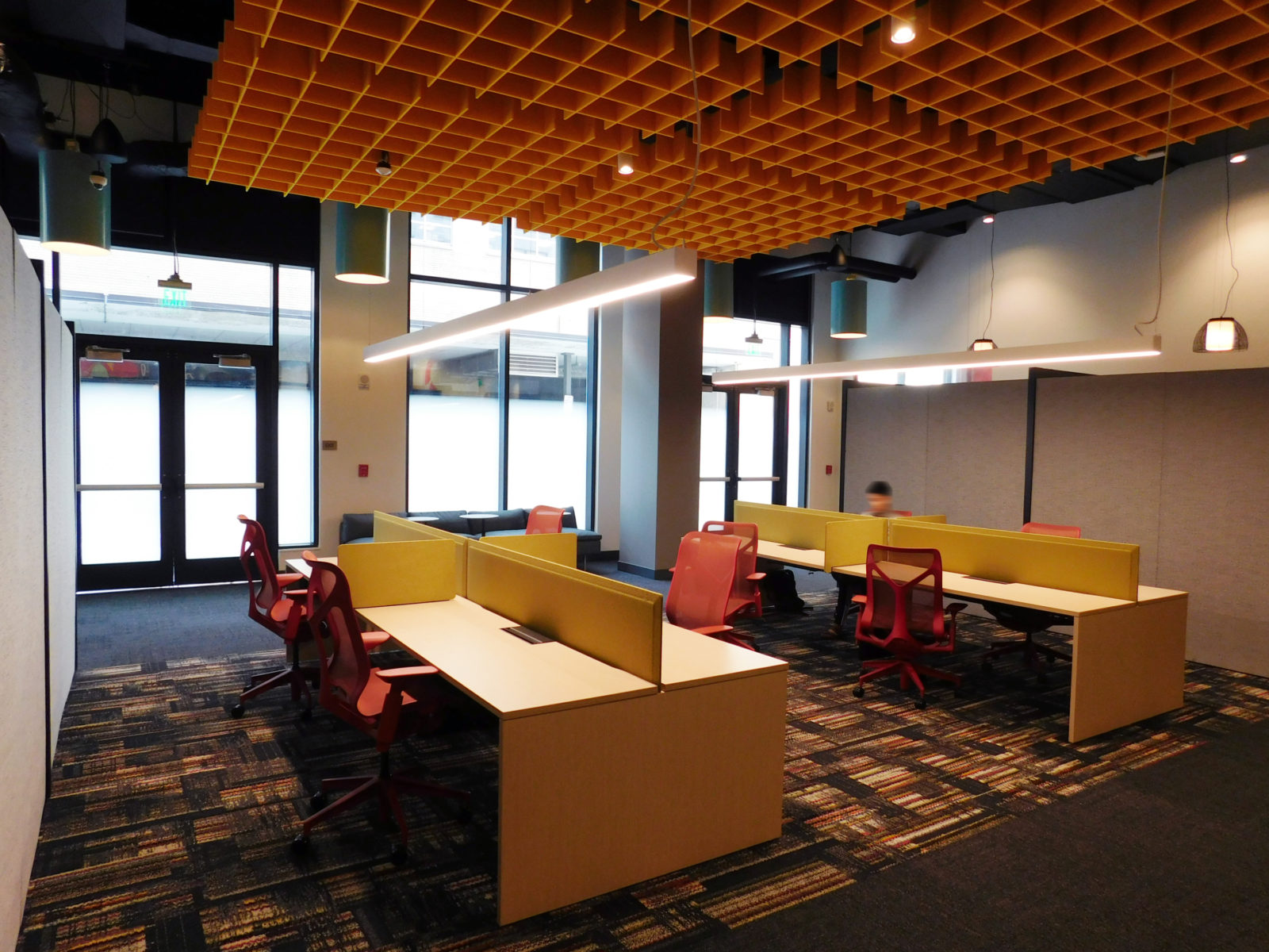 Two clusters of benching workstations with mustard yellow fabric screens on surface and red Herman Miller Cosm chairs at each spot. Acoustic panels above.