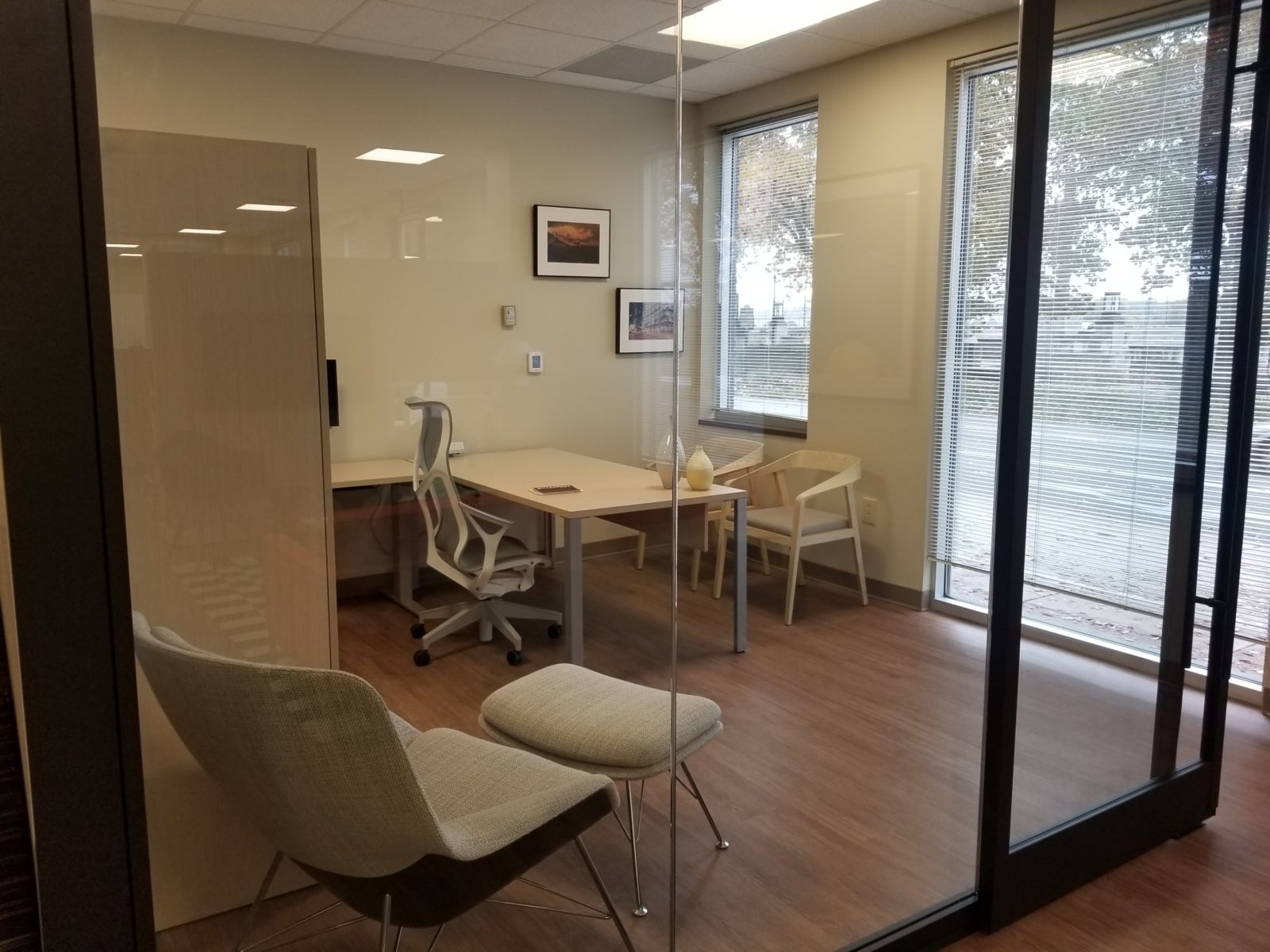 Glass DIRTT wall and door with black hardware and trim; inside room is a light woodgrain private office with Herman Miller Cosm chair and two guest chairs