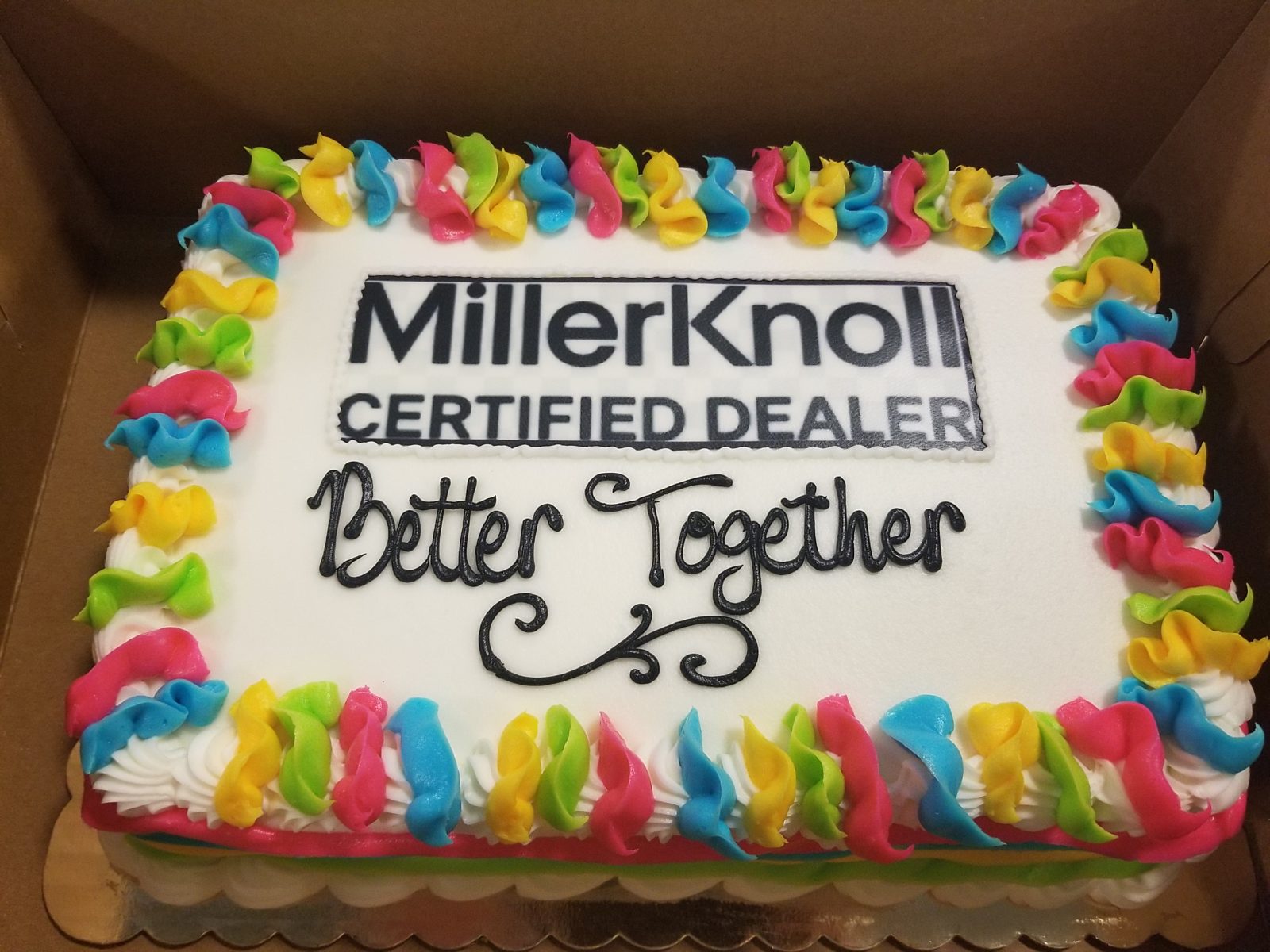 Custom cake that has the MillerKnoll Certified Dealer logo on it along with writing that says "Better Together" - edge is blue, yellow, green and pink