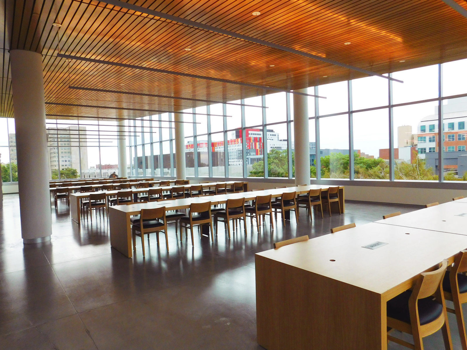 Open room with full windows on all sides, wooden slat ceiling and rows of long collaborative tables and chairs.
