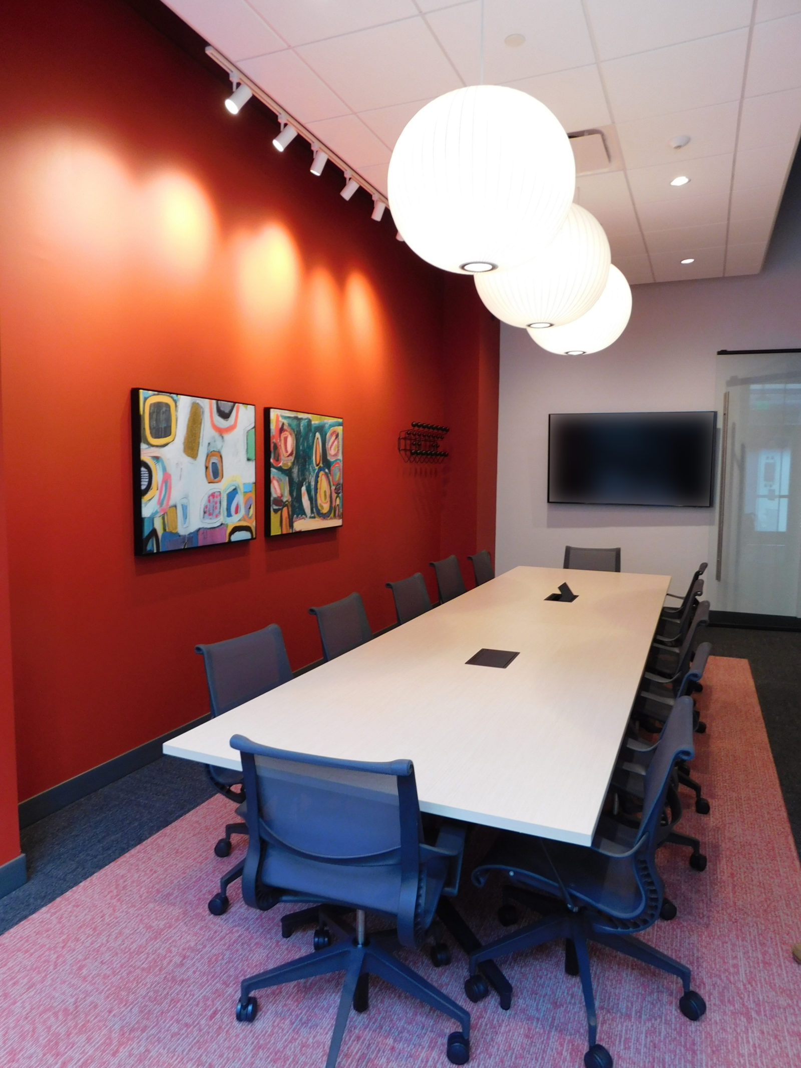 Conference room with white table and dark grey Herman Miller Setu chairs around. Red wall on left; 3 white bubble lamps above table.