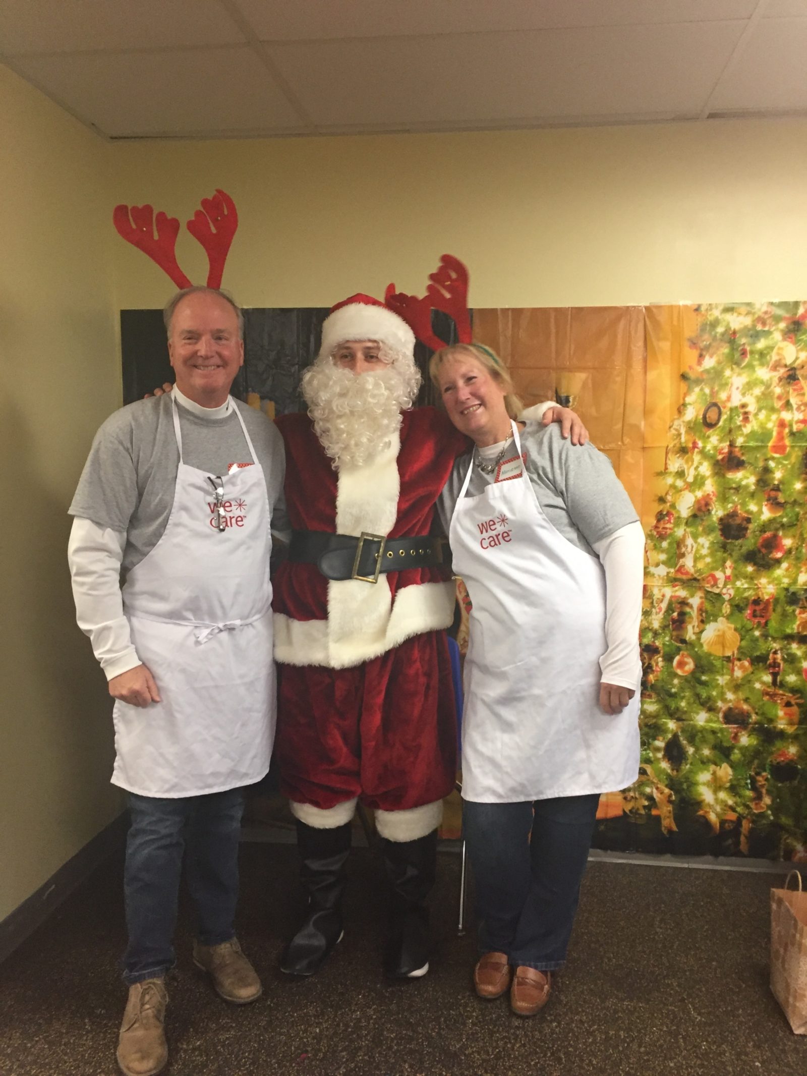 Santa Claus is in the middle of two people, one male and one female. They are wearing "We Care" aprons. There is a Christmas tree to the right side.
